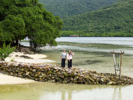Aerial view of two figures stand on a rocky breakwater gazing out at the shimmering turquoise water and lush green hills Bobby Beach Karimunjawa Central Java Indonesia.