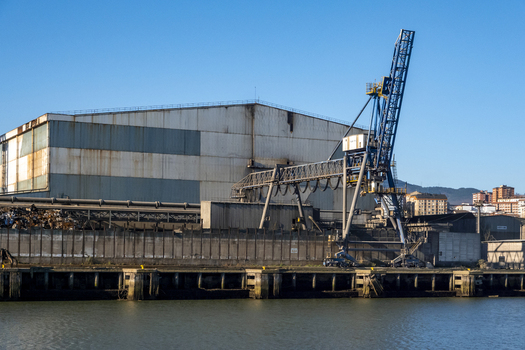 View of a large industrial warehouse and a blue harbor crane along the waterfront under a clear sky in Greater Bilbao Basque Country Spain. View of a large industrial warehouse and a blue harbor crane along the waterfront under a clear sky in Greater Bilbao Basque Country Spain.