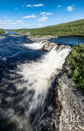 Aerial view of Pikefossen falls with rushing water cascading over rocky ledges surrounded by lush green vegetation under a clear blue sky in Kautokeino Finnmark Norway. Aerial view of Pikefossen falls with rushing water cascading over rocky ledges surrounded by lush green vegetation under a clear blue sky in Kautokeino Finnmark Norway.