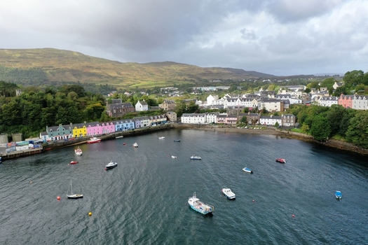 Aerial view of Portree Harbour with colorful houses lining the waterfront small boats anchored in the bay and rolling green hills under a cloudy sky in Portree Scotland United Kingdom.