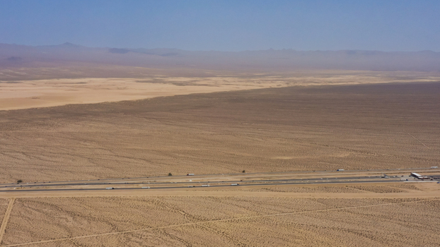 Aerial view of a highway stretching across the vast arid desert landscape with distant mountains under a clear sky in the United States.