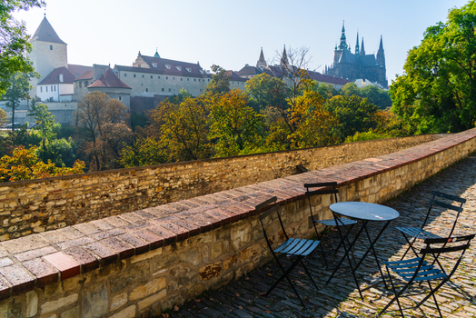 View of Prague Castle and St. Vitus Cathedral seen from a stone terrace with a metal table and chairs surrounded by autumn trees in Prague Prague Czechia.