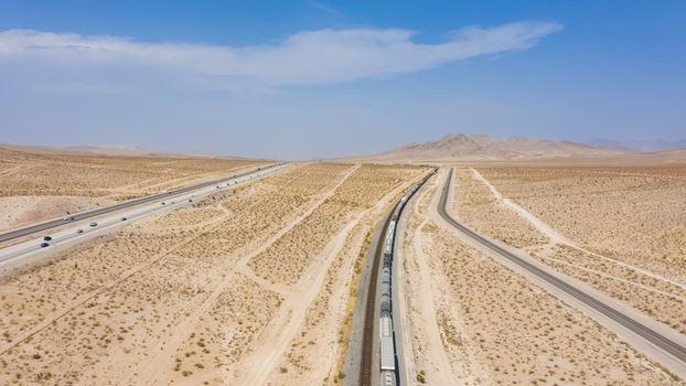 Aerial view of a freight train traveling on tracks alongside a highway through the arid desert landscape of the United States.