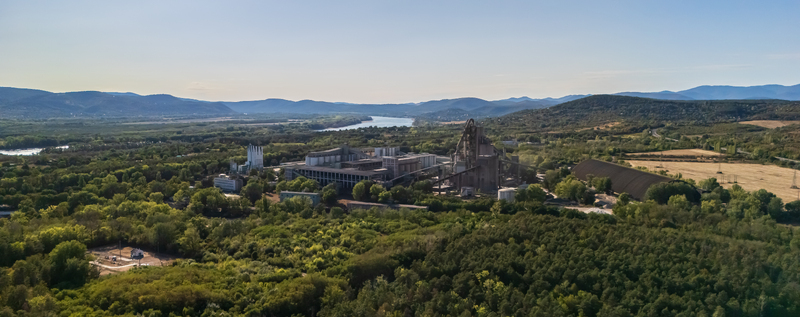 Aerial view of a cement manufacturing plant with silos and processing machinery beside the Danube River surrounded by forested hills and farmland in Vac Hungary during summer. Aerial view of a cement manufacturing plant with silos and processing machinery beside the Danube River surrounded by forested hills and farmland in Vac Hungary during summer.