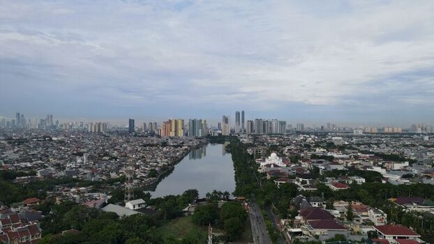 Aerial view of a canal flowing through a dense urban area with residential rooftops lush trees and high rise buildings under a cloudy sky North Jakarta Jakarta Indonesia. Aerial view of a canal flowing through a dense urban area with residential rooftops lush trees and high rise buildings under a cloudy sky North Jakarta Jakarta Indonesia.