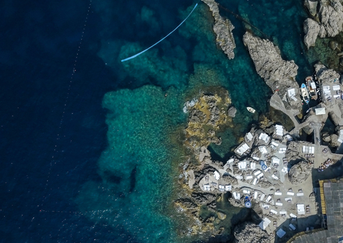 Aerial view of the Faraglioni rocky coastline featuring a beach club with white sunbeds and umbrellas by the turquoise sea in Capri Campania Italy.