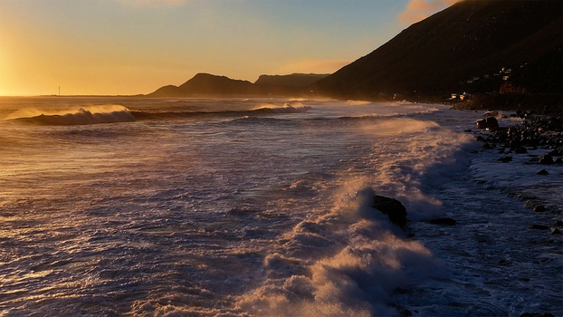 Aerial view of powerful waves crashing against a rocky coastline under a golden sunset with steep mountain slopes in Cape Town Western Cape South Africa. Aerial view of powerful waves crashing against a rocky coastline under a golden sunset with steep mountain slopes in Cape Town Western Cape South Africa.