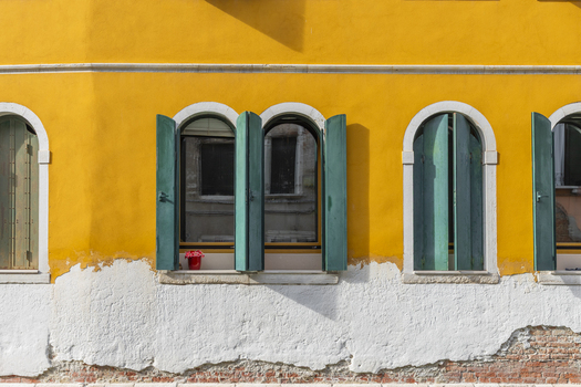 View of a vibrant yellow wall with weathered white plaster featuring arched windows with open green shutters and a small red flower pot in Venice Veneto Italy. View of a vibrant yellow wall with weathered white plaster featuring arched windows with open green shutters and a small red flower pot in Venice Veneto Italy.