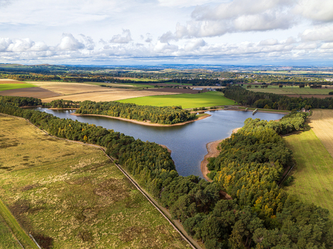 Aerial view of Harlaw Reservoir surrounded by lush green forests agricultural fields and rolling hills under a cloudy sky in Balerno Scotland United Kingdom.