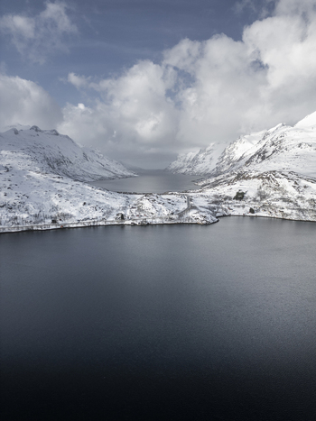 Aerial view of Grotfjord with snow capped mountains dark deep waters and a coastal road under a cloudy sky in Tromvik Troms Norway. Aerial view of Grotfjord with snow capped mountains dark deep waters and a coastal road under a cloudy sky in Tromvik Troms Norway.