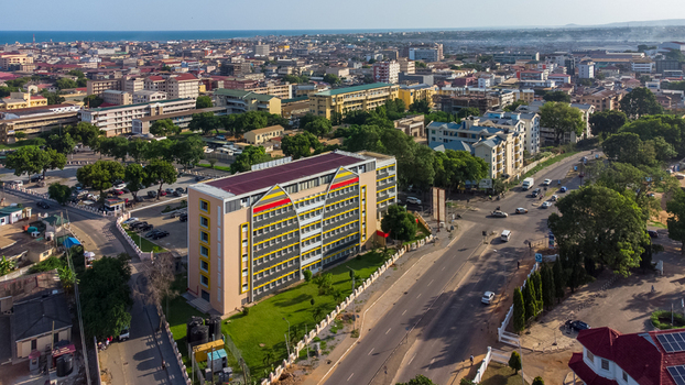 Accra Ghana 26 May 2023 Aerial view of a modern multi story office building with a distinctive yellow and red facade surrounded by urban streets green trees and the cityscape of Accra. Accra Ghana 26 May 2023 Aerial view of a modern multi story office building with a distinctive yellow and red facade surrounded by urban streets green trees and the cityscape of Accra.