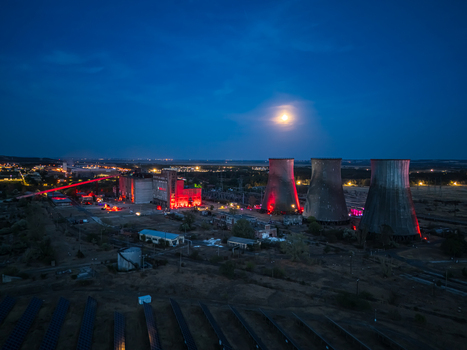 Aerial view of illuminated cooling towers and abandoned industrial buildings at a former thermal power plant during a festival in Inota Hungary with solar panels under a moonlit evening sky.