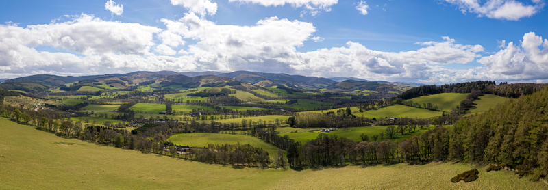 Aerial view of the rolling green hills and forests of Glentress featuring lush meadows and distant mountains under a bright blue sky in Glentress Scotland United Kingdom.