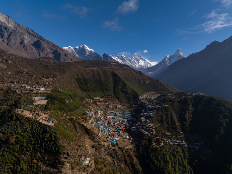 Aerial view of a mountain village nestled in the Himalayas with colorful rooftops surrounding pine forests and snow capped peaks under a clear blue sky in Nepal.