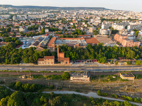 Aerial view of the historic industrial complex with a tall brick chimney and old factory buildings surrounded by lush trees near railway tracks in Varna Varna Bulgaria. Aerial view of the historic industrial complex with a tall brick chimney and old factory buildings surrounded by lush trees near railway tracks in Varna Varna Bulgaria.