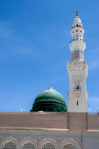 View of the iconic green dome and tall white minaret of Al Masjid e Nabawi against a clear blue sky in Madinah Al Madinah Province Saudi Arabia.
