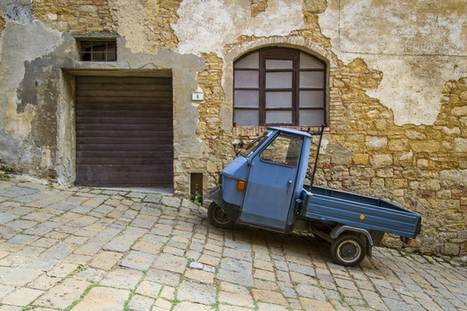 View of a blue Piaggio Ape parked on a stone street against a weathered stone building with peeling plaster creating a rustic scene Volterra Tuscany Italy. View of a blue Piaggio Ape parked on a stone street against a weathered stone building with peeling plaster creating a rustic scene Volterra Tuscany Italy.