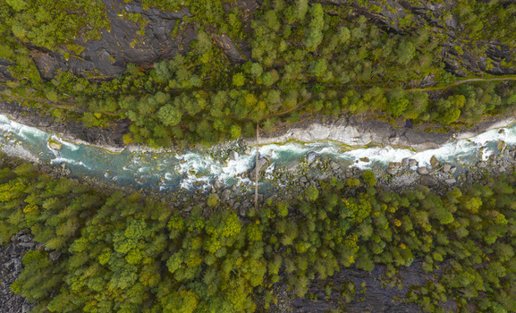 Aerial view of a turquoise river flowing through a rocky canyon and dense green forest with a small wooden bridge in Fjaera Vestland Norway. Aerial view of a turquoise river flowing through a rocky canyon and dense green forest with a small wooden bridge in Fjaera Vestland Norway.