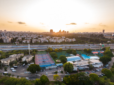Aerial view of a sports complex with tennis courts and a swimming pool surrounded by trees and urban buildings under a golden sunset sky in Tel Aviv Yafo Tel Aviv District Israel. Aerial view of a sports complex with tennis courts and a swimming pool surrounded by trees and urban buildings under a golden sunset sky in Tel Aviv Yafo Tel Aviv District Israel.
