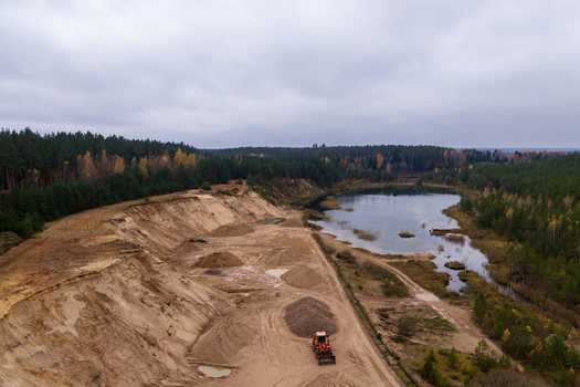 Aerial view of a sand quarry with an orange excavator piles of sand and a calm lake surrounded by dense autumn forest under a cloudy sky Nemencine Vilnius County Lithuania. Aerial view of a sand quarry with an orange excavator piles of sand and a calm lake surrounded by dense autumn forest under a cloudy sky Nemencine Vilnius County Lithuania.