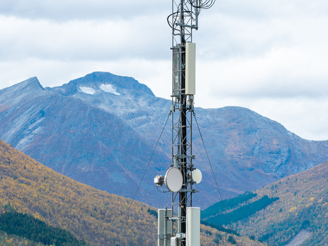 Aerial view of a telecommunications tower with various antennas and equipment set against a backdrop of rugged mountains and autumn colored forests in Fjord More og Romsdal Norway.