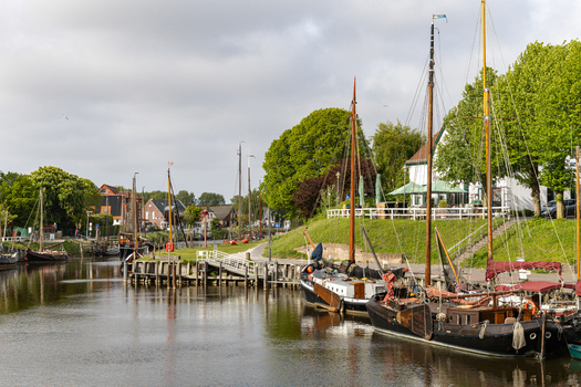View of Hafen Harlesiel with traditional sailing boats moored along the canal surrounded by lush green trees and historic buildings in Wittmund Lower Saxony Germany. View of Hafen Harlesiel with traditional sailing boats moored along the canal surrounded by lush green trees and historic buildings in Wittmund Lower Saxony Germany.
