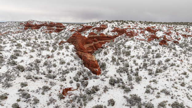 Aerial view of red sandstone rock formations and desert shrubs covered in a fresh layer of white snow under a cloudy sky in Page Arizona United States.