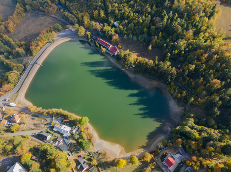 Aerial view of Velka Vindsachta lake surrounded by dense autumn forests winding roads and small residential buildings in Stiavnicke Bane Banskobystricky kraj Slovakia.