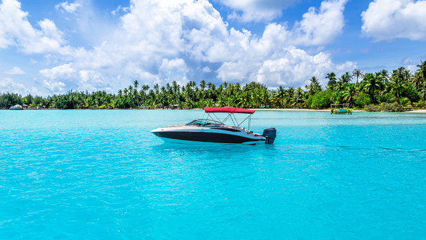 View of a motorboat with a red canopy floating on clear turquoise water with a lush palm fringed coastline in the background in Bora Bora Leeward Islands French Polynesia.