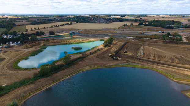 Aerial view of the Black Cat Roundabout construction site featuring blue lakes earthworks and heavy machinery in the rural landscape of Chawston England United Kingdom.