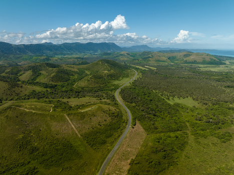 Aerial view of a winding road cutting through the lush green rolling hills and dense forests under a bright blue sky dotted with fluffy white clouds Noumea South Province New Caledonia.