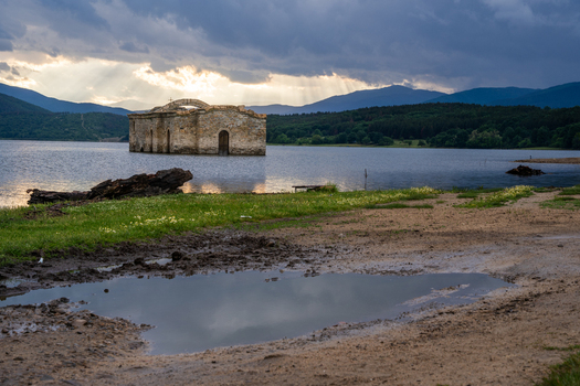 View of the submerged St. John of Rila church in a lake with dramatic sunbeams breaking through storm clouds over distant mountains and a muddy shoreline in Sliven Bulgaria. View of the submerged St. John of Rila church in a lake with dramatic sunbeams breaking through storm clouds over distant mountains and a muddy shoreline in Sliven Bulgaria.
