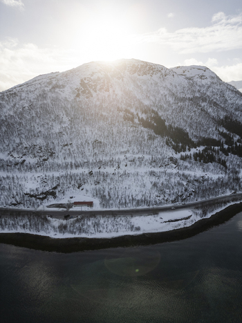 Aerial view of Grotfjord with a snow covered mountain peak a coastal road and the sun shining over the ridge in Tromvik Troms Norway. Aerial view of Grotfjord with a snow covered mountain peak a coastal road and the sun shining over the ridge in Tromvik Troms Norway.