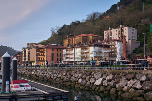 Pasai Donibane Spain 09 March 2026 View of the main harbor street with people at outdoor cafes along the stone waterfront under historic buildings and a forested hillside.