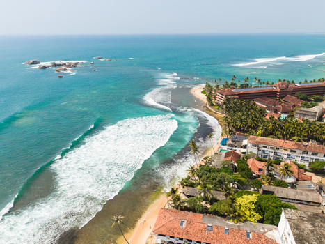 Aerial view of turquoise ocean waves crashing against a sandy beach lined with palm trees and coastal resorts in Hikkaduwa Southern Province Sri Lanka. Aerial view of turquoise ocean waves crashing against a sandy beach lined with palm trees and coastal resorts in Hikkaduwa Southern Province Sri Lanka.