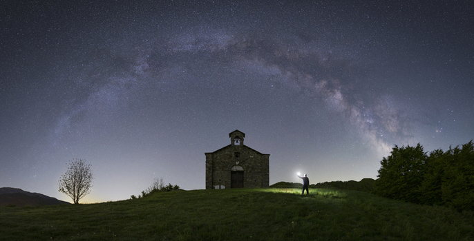 View of the stone chapel at Passo di Cirone under the Milky Way arch with a person holding a light on a grassy hill in Pontremoli Toscana Italy.