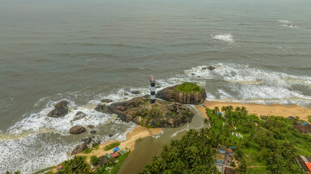 Aerial view of the iconic black and white striped Kapu Light House situated on a rocky outcrop surrounded by the Arabian Sea sandy beach and lush palm trees in Udupi Karnataka India.