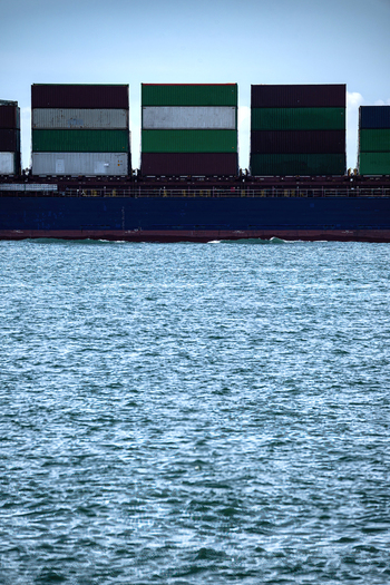 View of a large cargo ship carrying colorful stacked containers across the blue sea under an overcast sky.