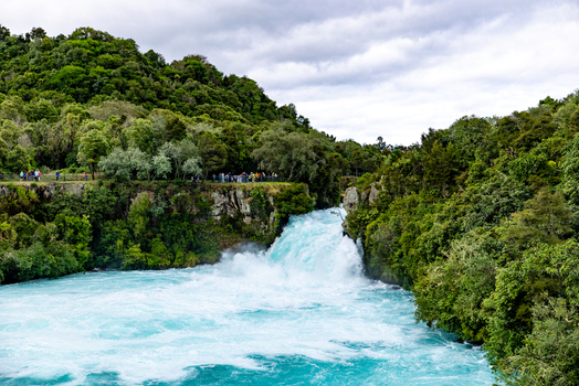 View of powerful turquoise waters thunder through a narrow gorge framed by lush green forests under a cloudy sky Huka Falls Waikato Region New Zealand. View of powerful turquoise waters thunder through a narrow gorge framed by lush green forests under a cloudy sky Huka Falls Waikato Region New Zealand.