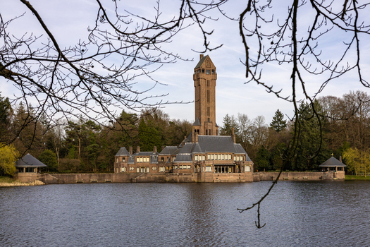 View of the Country Residence Museum Jachthuis Sint Hubertus with its tall brick tower overlooking a calm lake and dense forest in Hoenderloo Gelderland Netherlands.