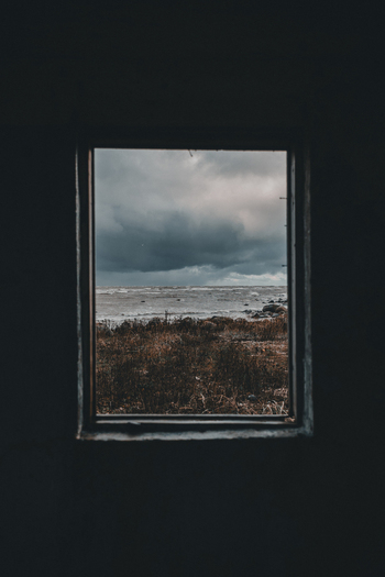 View of the stormy sea and rocky coastline of Vormsi island framed by a dark interior window with dramatic dark clouds and dry coastal vegetation in Laane County Estonia.