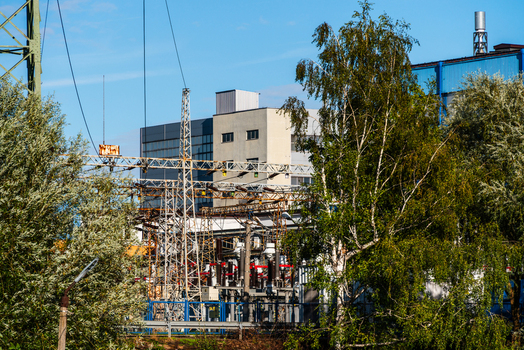 View of an electrical substation with high voltage power lines and metal structures set against an industrial building and lush green trees in Poland.