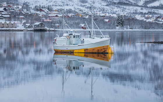 View of a white and yellow fishing boat anchored in calm water with reflections and a snow covered village in Tromso Troms Norway. View of a white and yellow fishing boat anchored in calm water with reflections and a snow covered village in Tromso Troms Norway.