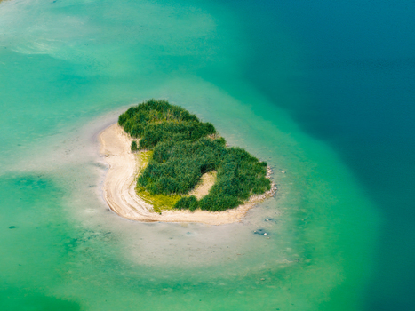 Aerial view of a small green island with lush vegetation and a sandy shore situated in a post industrial lake with vibrant turquoise water in Poland.