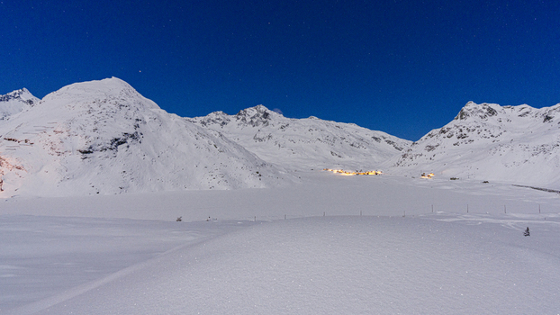 View of snow covered alpine peaks under a starry night sky with the distant lights of a mountain village in Madesimo Lombardy Italy. View of snow covered alpine peaks under a starry night sky with the distant lights of a mountain village in Madesimo Lombardy Italy.