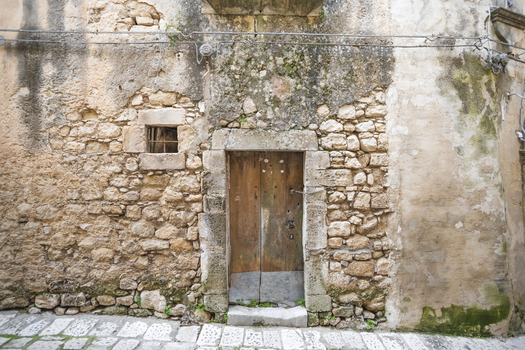 View of a rustic wooden door set in an ancient stone wall with a small barred window and mossy textures in Ragusa Sicily Italy.