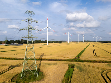 Aerial view of wind turbines in a golden wheat field with a large electricity pylon in the foreground under a blue sky. Symbolizing Sustainable Energy Transition and Infrastructure Aerial view of wind turbines in a golden wheat field with a large electricity pylon in the foreground under a blue sky. Symbolizing Sustainable Energy Transition and Infrastructure