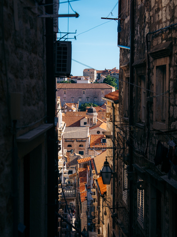 View of a narrow stone alleyway looking towards sunlit terracotta rooftops and a historic church dome under a clear blue sky in Dubrovnik Dubrovnik Neretva County Croatia. View of a narrow stone alleyway looking towards sunlit terracotta rooftops and a historic church dome under a clear blue sky in Dubrovnik Dubrovnik Neretva County Croatia.