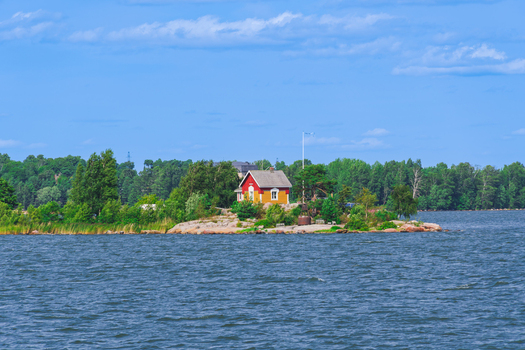 View of a small red and yellow wooden house on a rocky island surrounded by calm waters with a lush forest in the background in Helsinki Uusimaa Finland.