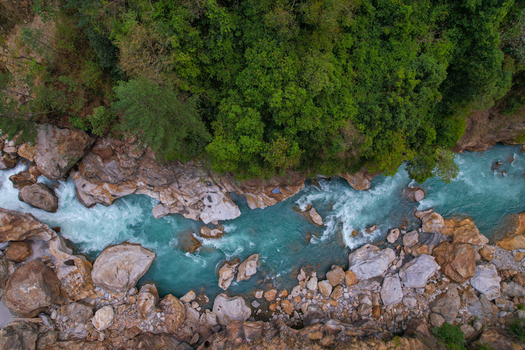 Aerial view of a turquoise river with white rapids flowing through a rocky gorge surrounded by a lush green forest in Nepal.